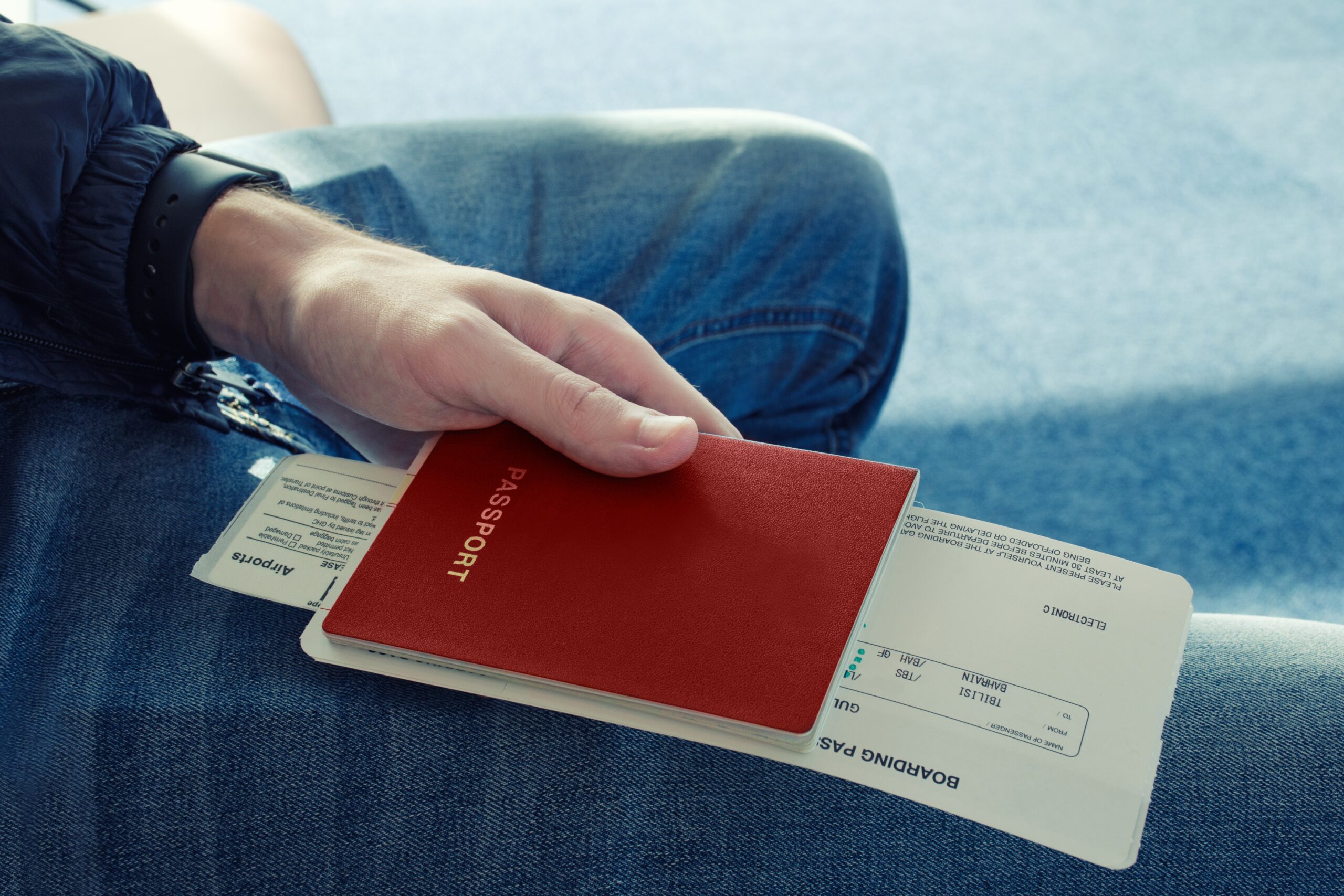 Man in blue jeans sits and holds in his hand passport of red color with tickets to plane. Сlose up. Top view.