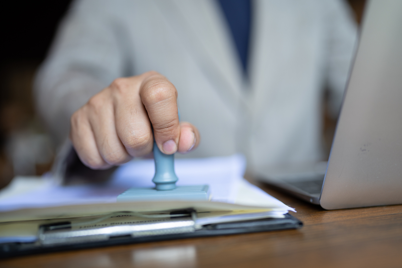 Close-up of a person's hand stamping with approved stamp on certificate document public paper at desk, notary or business people work from home, isolated for coronavirus COVID-19 protection
