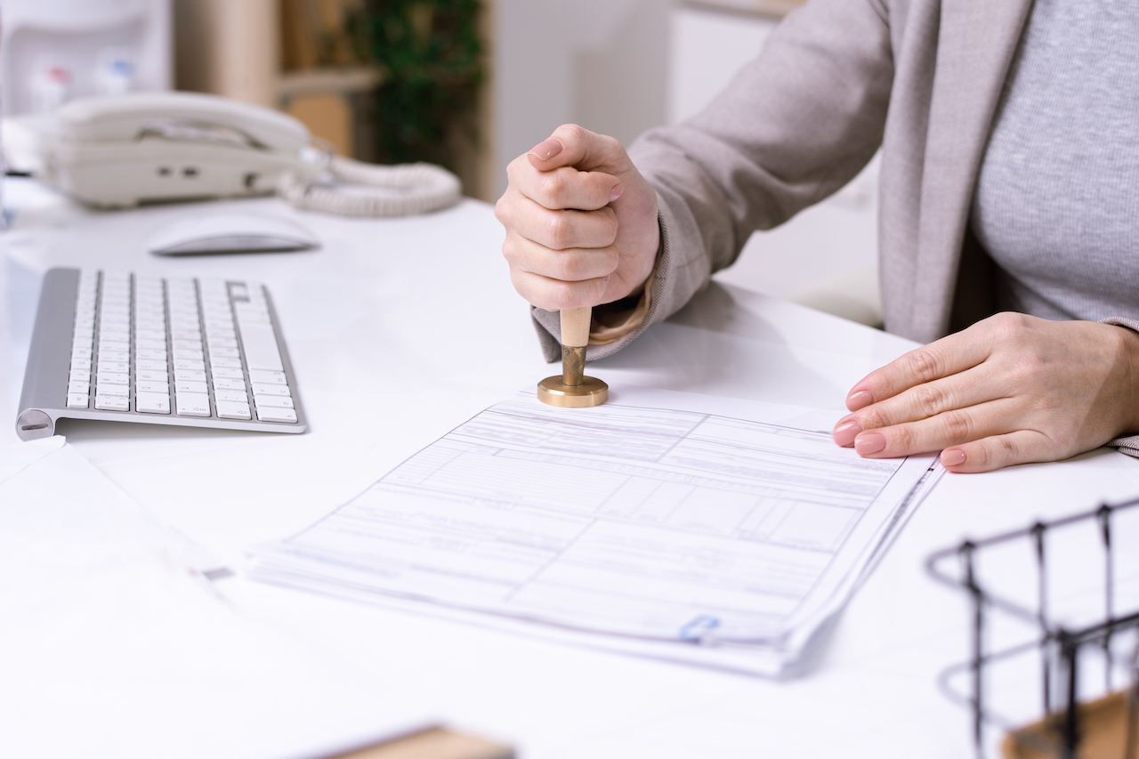 Hands of young female office worker sitting by desk and putting seal on financial document before sending it to client