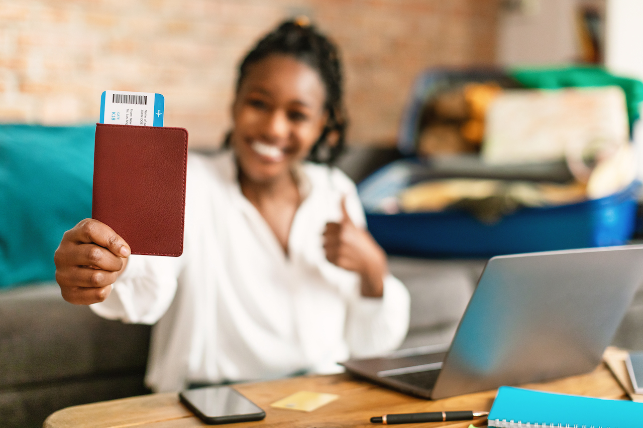 Travelling, vacation, tourism concept. Happy black woman showing passport with flight tickets and thumb up, smiling at camera, preparing for trip and packing suitcase